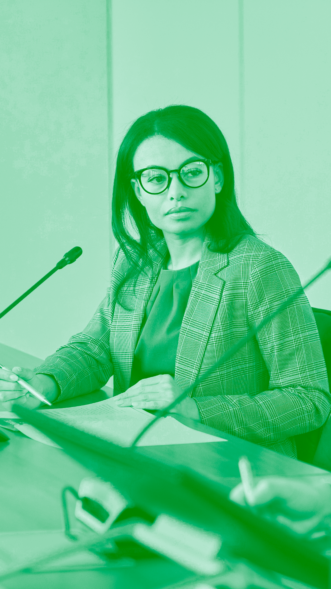 Mulher negra usando blusa branca, blazer e óculos de grau, sentada em uma mesa com um microfone à frente dela, olhando para a esquerda, para outra pessoa que também usa um microfone. Não é possívevl ver a outra pessoa na imagem.